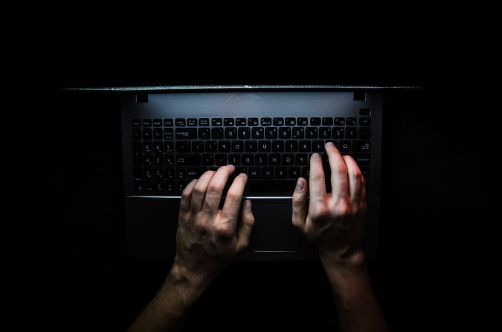 A person sitting in the dark types on a laptop, their hands illuminated.