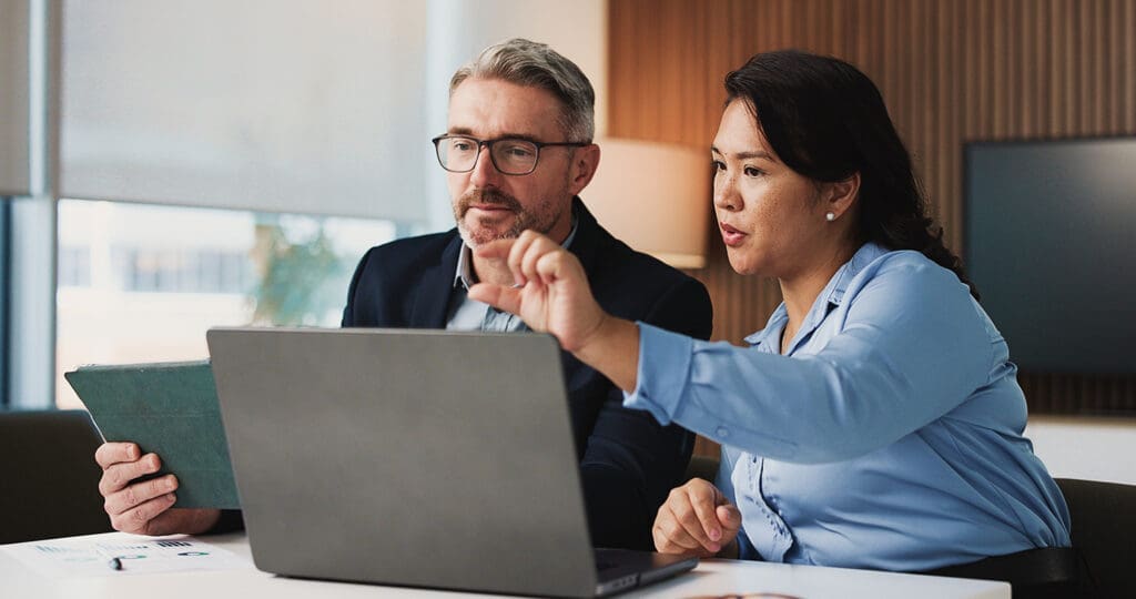 A man and a woman talking in front of a laptop screen in a business office.